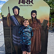 Emberlynn is registered to the contest to win money with this photo: child, children, face_cutout, smile, outdoor, daylight, clouds, sky, board, costume, play, fun, park, casual_clothing, shorts, sneakers, pavement, tree, greenery, signage