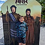 child, children, face_cutout, smile, outdoor, daylight, clouds, sky, board, costume, play, fun, park, casual_clothing, shorts, sneakers, pavement, tree, greenery, signage