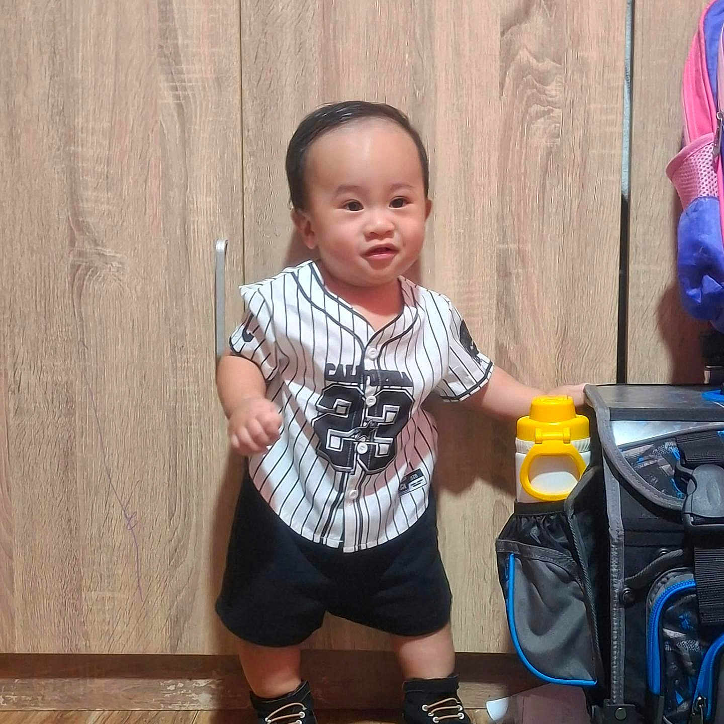 Zyron is registered to the contest to win money with this photo: toddler, child, standing, portrait, smiling, striped_jersey, baseball_jersey, black_shorts, black_shoes, wooden_wardrobe, indoors, wooden_floor, backpack, bottle, clothing, short_hair, face, person, home, photograph