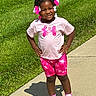 child, girl, smile, outdoor, sidewalk, grass, sunlight, pink_clothing, shorts, tshirt, sneakers, hair_accessories, happy, standing, casual, daytime, cute, confident, person, portrait