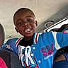 child, boy, smiling, portrait, selfie, car, vehicle, passenger, backseat, indoors, jacket, blue_clothing, varsity_jacket, t_shirt, short_hair, teeth, happy, sitting, automobile, daylight