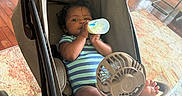 Dalton is registered to the contest to win money with this photo: baby, bottle, stroller, fan, indoor, wooden_floor, rug, onesie, child, infant, cooling_fan, seated, drink, baby_clothes, person, foot, hand, window, natural_light, relaxing