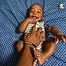 baby, background, bed, blue_sheet, child, fingers, foot, hand, happy, infant, joyful, laughing, onesie, patterned_sheet, person, playing, skin, smiling, striped_clothing, tickling