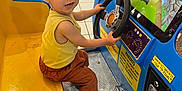 Thiméo a rejoint le concours — aidez-le/la à gagner de superbes lots ! child, toddler, arcade_game, kiddie_ride, steering_wheel, mall, tiled_floor, yellow_seat, blue_console, smiling, brown_hair, sneakers, casual_clothing, pants, playing, gaming, coin_slot, screen, indoor, family_fun
