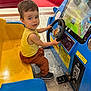 child, toddler, arcade_game, kiddie_ride, steering_wheel, mall, tiled_floor, yellow_seat, blue_console, smiling, brown_hair, sneakers, casual_clothing, pants, playing, gaming, coin_slot, screen, indoor, family_fun