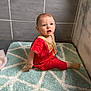 baby, barefoot, candid, casual, child, curious_expression, floor, geometric_pattern, gold_embroidery, home_interior, indoor, light, person, plush_rug, portrait, red_clothing, rug, sitting, tile_wall, young_child