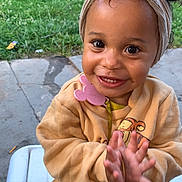Naziyah is registered to the contest to win money with this photo: toddler, child, smiling, clapping, head_wrap, outdoor, bench, yellow_clothing, pacifier, happy, cute, baby, person, face, skin, eyes, hands, grass, concrete, daylight