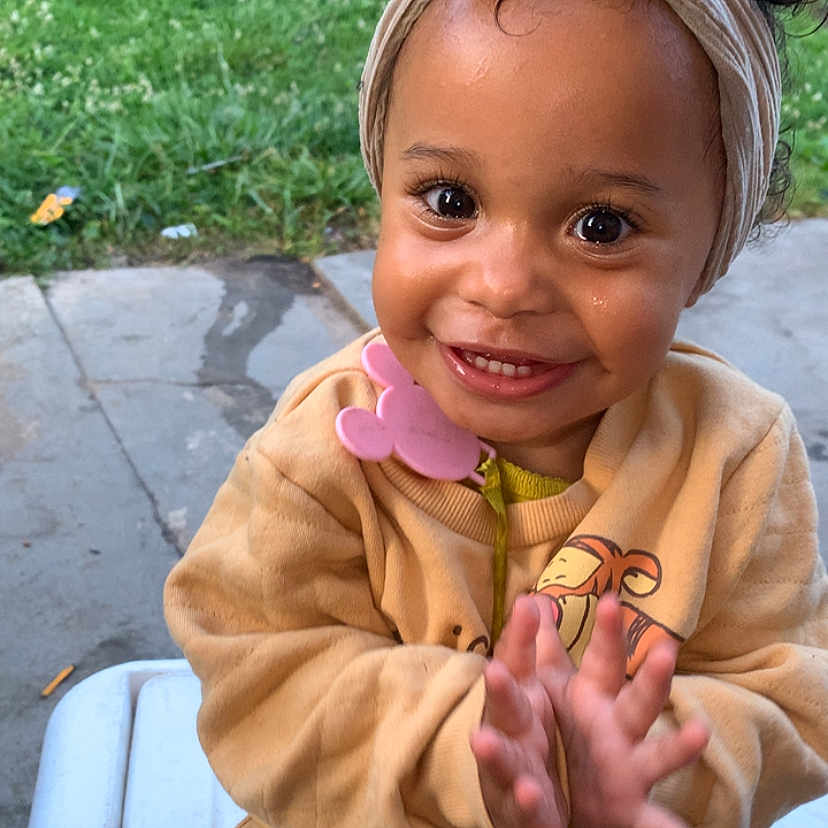 Naziyah is registered to the contest to win money with this photo: toddler, child, smiling, clapping, head_wrap, outdoor, bench, yellow_clothing, pacifier, happy, cute, baby, person, face, skin, eyes, hands, grass, concrete, daylight