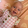 baby, smiling, infant, child, blanket, rainbow_pattern, pink_clothing, checkered_pattern, floral_pattern, wooden_buttons, happy, face, portrait, lying_down, cute, skin, person, indoor, closeup, playful