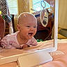 baby, mirror, reflection, smile, face, onesie, pink, patterned_clothing, infant, happy, indoor, bed, blanket, toy, home, window, light, furniture, curtains, playtime