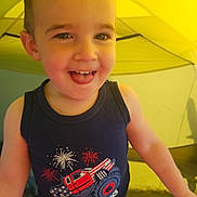 Logan is registered to the contest to win money with this photo: toddler, child, smiling, tank_top, monster_truck, fireworks, indoor, tent, yellow, cute, happy, face, portrait, clothing, arms, baby, person, young_child, fun, playful