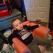 Kason joined the competition — help win amazing prizes! toddler, child, pink_armchair, black_shirt, text_on_clothing, green_pants, indoor, door, toy, clutter, floor, cabinet, fan, person, furniture, expression, room, casual, seated, curious