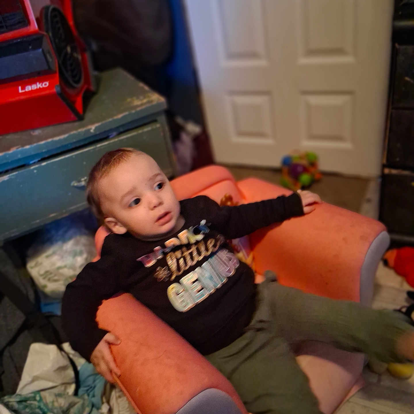 Kason joined the competition — help win amazing prizes! black_shirt, cabinet, casual, child, clutter, curious, door, expression, fan, floor, furniture, green_pants, indoor, person, pink_armchair, room, seated, text_on_clothing, toddler, toy