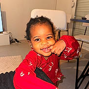 Fatoumata-Zahra participe au concours pour gagner de l'argent avec cette photo : child, toddler, smile, red_clothing, long_sleeve, finger_in_mouth, hand, eyes, earrings, hair, indoor, chair, table, rug, floor, portrait, happy, flash_lighting, home_interior, person