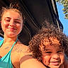 woman, child, smile, tattoo, curly_hair, sunlight, outdoor, blue_sky, tree, vehicle, portrait, happy, skin, face, shoulder, daylight, casual, people, nature, summer