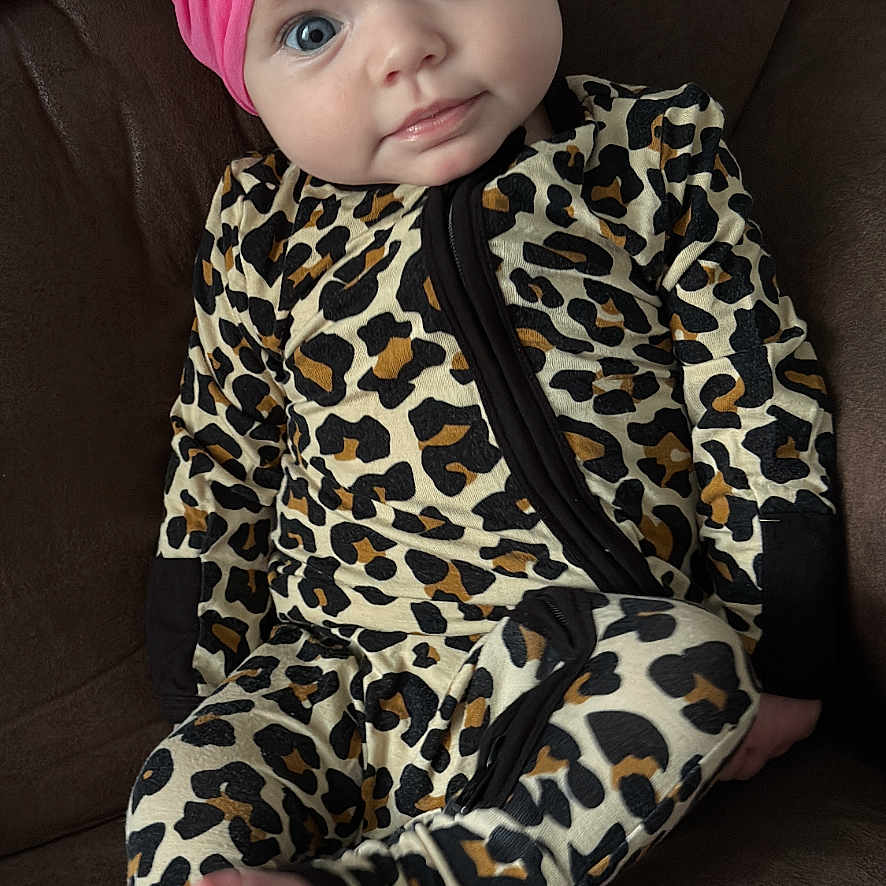 Riley Faye is registered to the contest to win money with this photo: baby, infant, headband, pink_bow, leopard_print, onesie, couch, brown_furniture, barefoot, foot, face, blue_eyes, curious_expression, seated, indoors, child, cute, portrait, young_child, comfortable