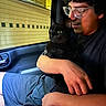 person, black_cat, glasses, car_interior, night, window, arm, seatbelt, shorts, hand, face, animal, pet, holding, looking, indoors, closeup, human, seated, comfort