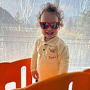 Lorenzo participe au concours pour gagner de l'argent avec cette photo : child, toddler, sunglasses, smile, curly_hair, tigger_shirt, long_sleeve_shirt, orange_playpen, play_fence, indoor, backlight, curtain, window, reflection, standing, happy, teeth, home, portrait, toy