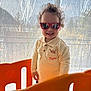 child, toddler, sunglasses, smile, curly_hair, tigger_shirt, long_sleeve_shirt, orange_playpen, play_fence, indoor, backlight, curtain, window, reflection, standing, happy, teeth, home, portrait, toy
