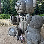 Luna is registered to the contest to win money with this photo: dog, french_bulldog, puppy, statue, sculpture, outdoor, garden, park, stone, leash, harness, pink, sitting, standing, animal, pet, small_dog, giant_dog, brick_wall, greenery