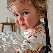 Hendrix is registered to the contest to win money with this photo: toddler, child, curly_hair, giraffe_pattern, onesie, bedroom, soft_lighting, wooden_stool, jars, bed, blanket, portrait, smile, face, indoor, cozy, cute, person, young_child, curious
