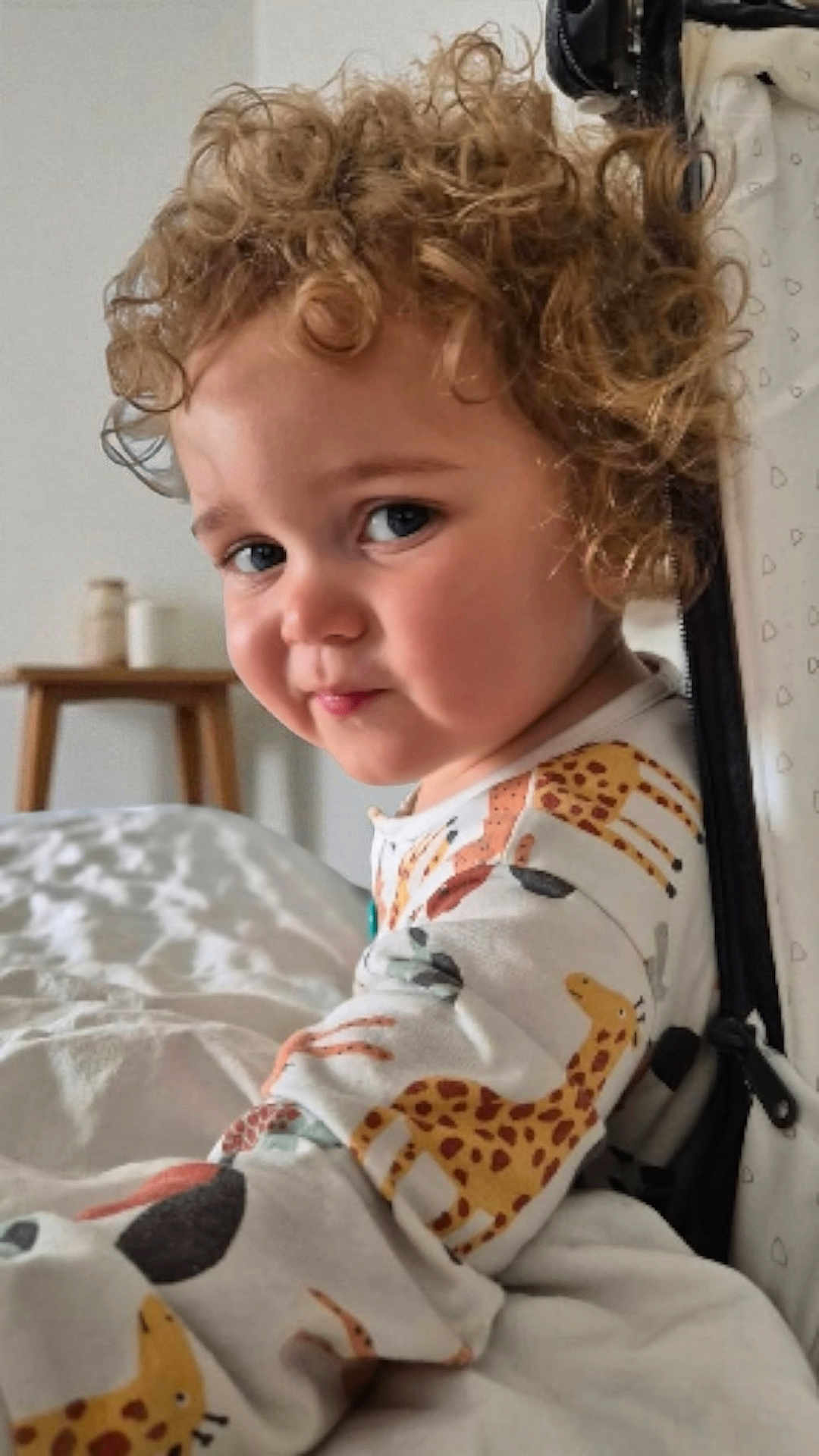Hendrix is registered to the contest to win money with this photo: toddler, child, curly_hair, giraffe_pattern, onesie, bedroom, soft_lighting, wooden_stool, jars, bed, blanket, portrait, smile, face, indoor, cozy, cute, person, young_child, curious