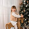 Kiara participe au concours pour gagner de l'argent avec cette photo : blonde_hair, child, christmas_tree, cozy, curtain, faux_fur_vest, festive, girl, handcrafted_toy, holiday_decor, indoor, lights, ornaments, pinecones, playful, portrait, seasonal_decor, smile, white_tights, wooden_rocking_horse