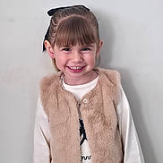 Kiara a rejoint le concours — aidez-le/la à gagner de superbes lots ! bangs, bow, child, cute, earrings, fashion, fur_vest, girl, hair, happy, indoor, long_sleeves, looking_at_camera, plain_wall, portrait, smile, standing, teeth, white_shirt, youth