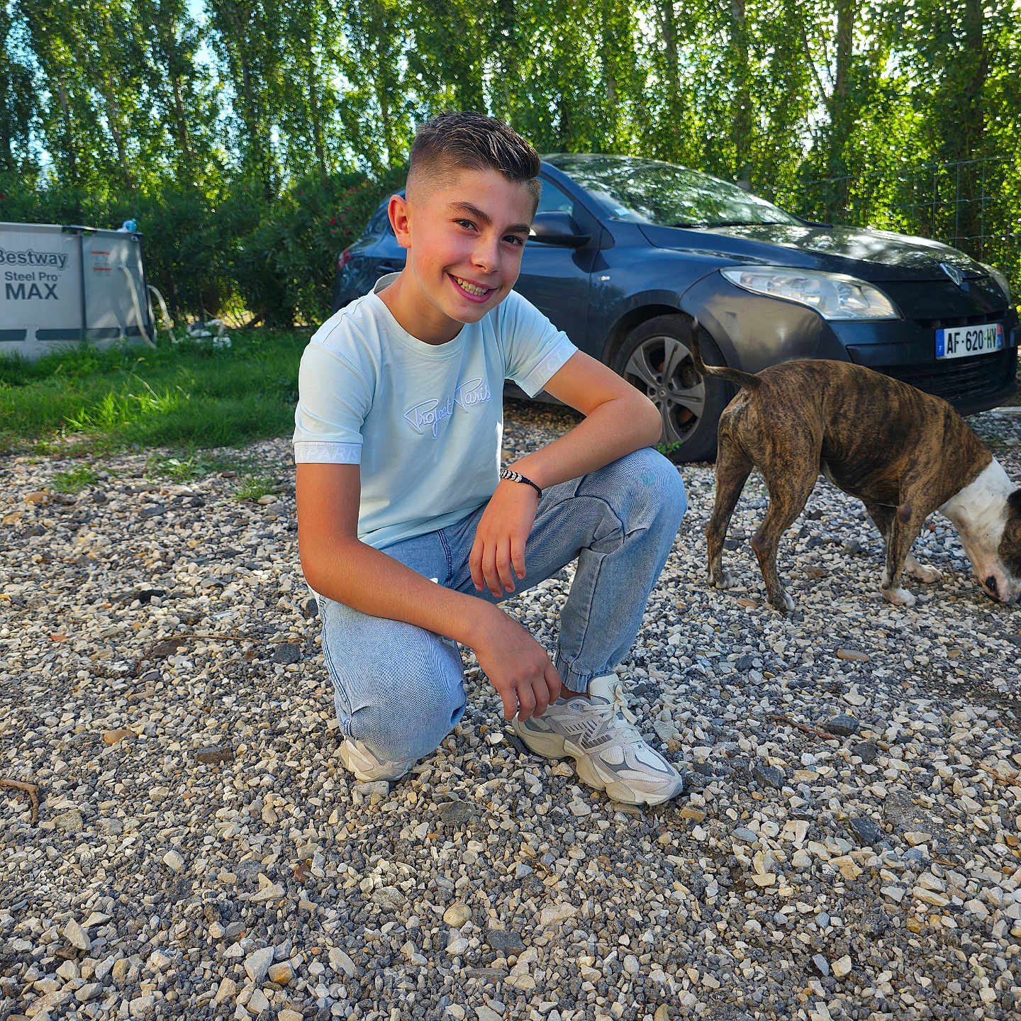 Edzio a rejoint le concours — aidez-le/la à gagner de superbes lots ! boy, brindle_dog, car, child, crouching, dog, gravel, greenery, happy, jeans, outdoors, parked_car, pet, portrait, smile, sneakers, sunlight, trees, tshirt, wristwatch