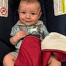 baby, infant, car_seat, chicco, blanket, smile, face, hand, clothing, safety_harness, red_blanket, blue_car_seat, warning_label, child, cute, young, indoor, seat, person, portrait