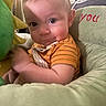 Beau is registered to the contest to win money with this photo: baby, child, face, smile, cushion, plush_toy, orange_shirt, indoor, close_up, person, infant, cute, happy, seated, soft_light, cozy, portrait, young_child, furniture, home