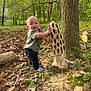 Riot is registered to the contest to win money with this photo: baby, child, outdoor, forest, tree, wood, carving, sculpture, leaves, nature, playful, curious, smiling, standing, greenery, trunk, sunlight, park, grass, casual_clothing