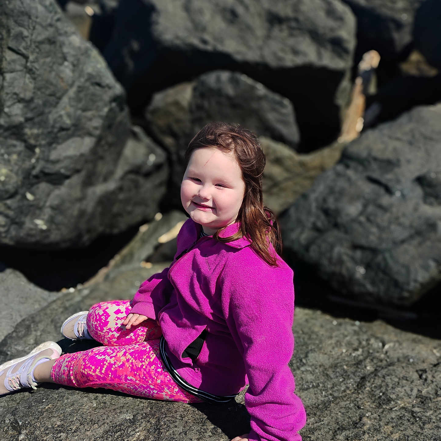 Rubie Jean is registered to the contest to win money with this photo: blue_sky, casual_clothing, child, daylight, hair, happy, nature, outdoor, patterned_leggings, person, pink_jacket, portrait, relaxing, rocks, scenery, shoes, sitting, smile, sunny, young