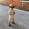 child, toddler, outdoor, sidewalk, brick_wall, building, sunglasses, sun_hat, striped_clothing, romper, sandals, toy, standing, portrait, urban, daylight, fashion, cute, person, street