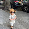 child, toddler, outdoor, sidewalk, city, street, sunglasses, sun_hat, striped_clothing, shirt, shorts, sandals, walking, standing, tree, parked_cars, urban, sunlight, portrait, fashion