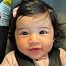 baby, infant, child, car_seat, seatbelt, harness, portrait, face, wide_eyes, looking_at_camera, indoors, vehicle, passenger, pink_clothing, hair_bow, dark_hair, sitting, close_up, safety_label, photography