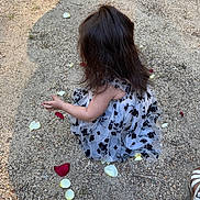 Miyrah is registered to the contest to win money with this photo: child, dress, flower_petals, gravel, ground, long_hair, outdoor, person, reaching_hand, sitting, white_dress, black_flower_pattern, footwear, sandals, nature, sunlight, shadow, texture, young_child, back_view