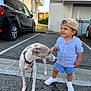 child, toddler, dog, parking_lot, car, cap, shorts, sneakers, sidewalk, leash, pet, outdoor, building, curious, smiling, striped_shirt, casual_clothing, daylight, person, animal