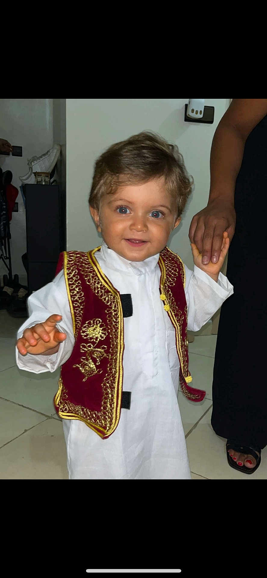 Lyam participe au concours pour gagner de l'argent avec cette photo : toddler, child, smiling, traditional_clothing, vest, white_outfit, indoor, holding_hand, person, blue_eyes, short_hair, floor, adult_hand, footwear, tile_floor, portrait, cute, happy, young_child, people