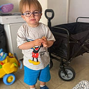 Noam a rejoint le concours — aidez-le/la à gagner de superbes lots ! blue_shoes, blue_shorts, cart, casual_clothing, child, floor, furniture, glasses, indoor, mickey_mouse, person, serious_expression, shoes, shorts, snack, standing, toy, tshirt, wagon, young_child