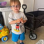 blue_shoes, blue_shorts, cart, casual_clothing, child, floor, furniture, glasses, indoor, mickey_mouse, person, serious_expression, shoes, shorts, snack, standing, toy, tshirt, wagon, young_child