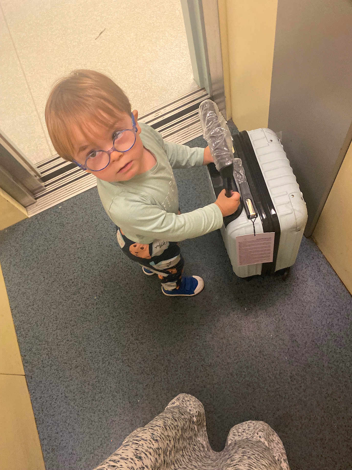 Noam a rejoint le concours — aidez-le/la à gagner de superbes lots ! child, glasses, suitcase, floor, elevator, door, plastic_wrap, blue, standing, looking_up, person, footwear, carpet, handle, travel, young_child, indoor, curious, round_glasses, clothing