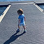 blue_shirt, casual_clothing, child, daytime, dock, glasses, metal_surface, nature, outdoor, person, river, shadow, sky, summer, sunlight, toddler, trees, walking, water, white_shorts