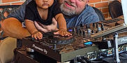 Ember is registered to the contest to win money with this photo: baby, child, man, dj_mixer, music_equipment, headband, red_bow, table, brick_wall, smile, glasses, technology, indoor, person, adult, device, hands, black_clothing, casual, footwear