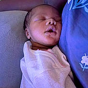 Ember is registered to the contest to win money with this photo: baby, newborn, sleeping, person, purple_shirt, floral_pattern, couch, blanket, close_up, indoor, rest, comfort, peaceful, face, head, infant, soft_lighting, portrait, tender_moment, relaxation