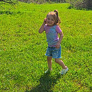 Kyleigh is registered to the contest to win money with this photo: child, girl, smiling, grass, outdoor, nature, greenery, tree, sunlight, denim_shorts, tie_dye_shirt, casual_clothing, happy, playful, standing, footwear, shadow, daylight, field, portrait