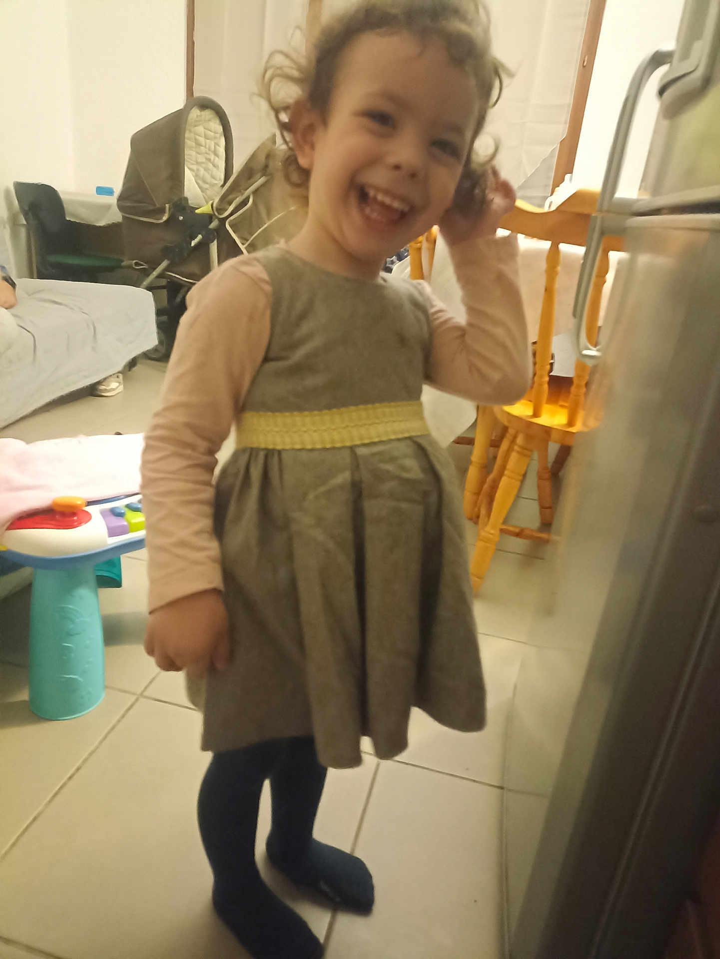 Mathilde Loureiro participe au concours pour gagner de l'argent avec cette photo : child, toddler, smiling, curly_hair, dress, indoor, floor, tile, toy, stroller, chair, table, furniture, happy, person, clothing, long_sleeve, socks, home, living_room
