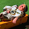 newborn, baby, infant, sleeping, portrait, outdoor, grass, sunlight, hat, cap, sports_theme, medallion, necklace, beads, onesie, clothing, socks, orange_box, seated, face