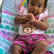 Zanovah is registered to the contest to win money with this photo: baby, child, infant, portrait, sitting, bouncer, baby_chair, smiling, looking_at_camera, hands_clasped, curly_hair, pink_clothing, plaid_shorts, socks, teddy_bear, colorful_background, outdoor, garden, sunlight, family_photo