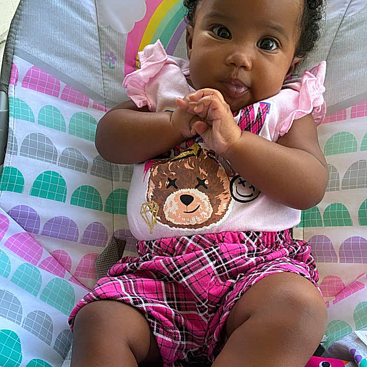 Zanovah is registered to the contest to win money with this photo: baby, child, infant, portrait, sitting, bouncer, baby_chair, smiling, looking_at_camera, hands_clasped, curly_hair, pink_clothing, plaid_shorts, socks, teddy_bear, colorful_background, outdoor, garden, sunlight, family_photo